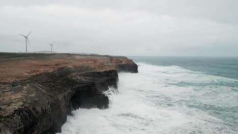 Slow-panning-aerial-shot-of-ocean-water-hitting-the-rocks-beside-a-wind-farm-in-Cape-Bridgewater,-Australia