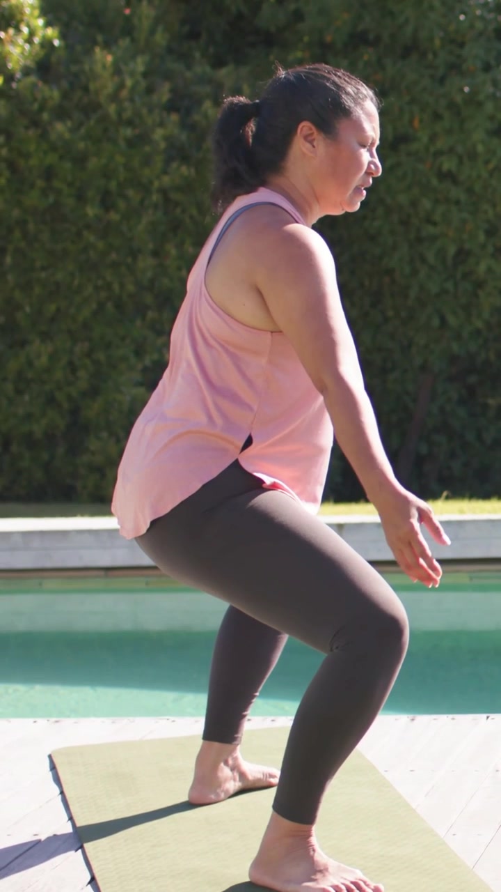 Video verticale: Una donna con top rosa inizia uno squat basso, facendo esercizi per la salute su un tappetino verde a bordo piscina.