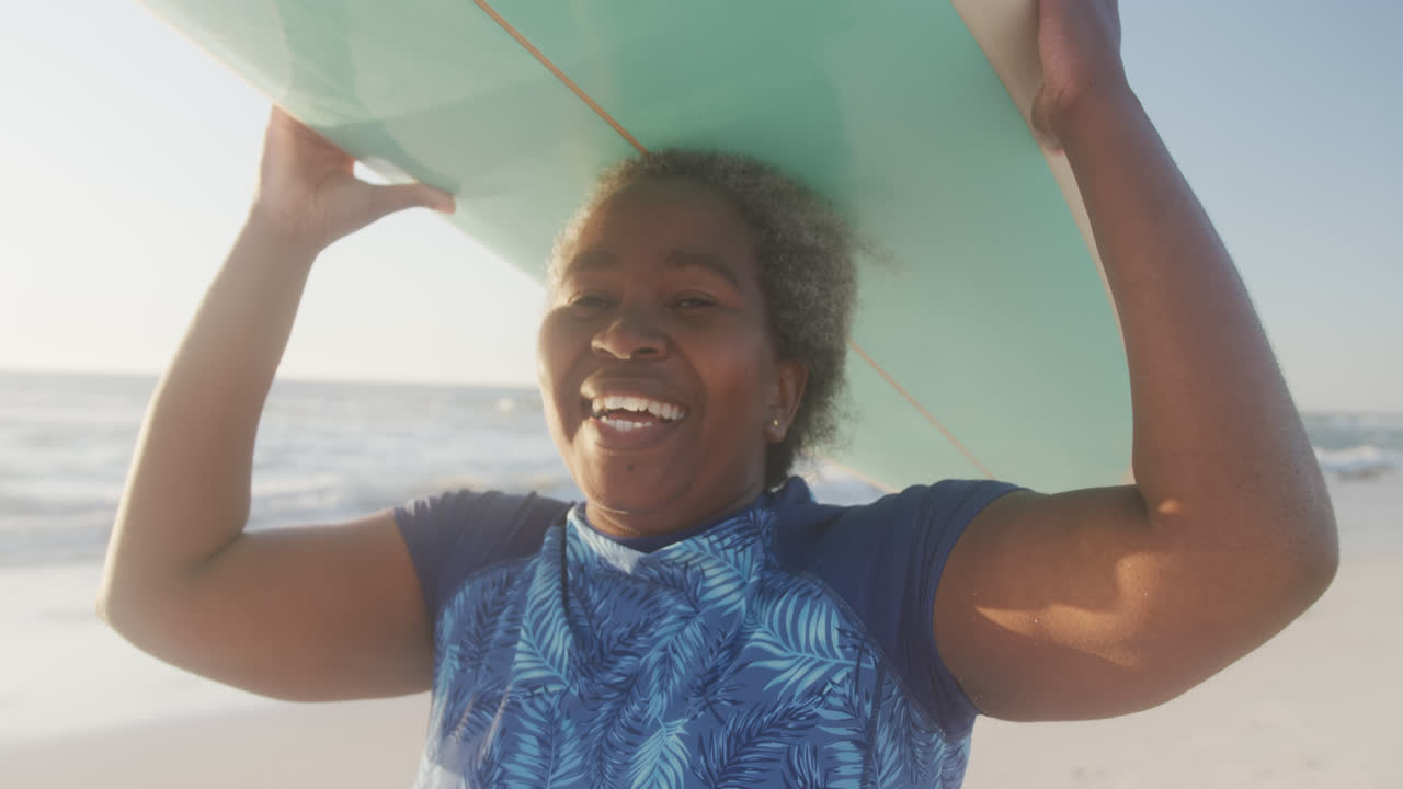 retrato de una feliz mujer afroamericana senior sosteniendo una tabla de surf en la playa, en cámara lenta