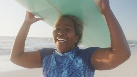 portrait of happy senior african american woman holding surfboard at beach, in slow motion