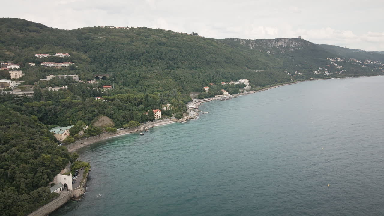 Un drone 4K UHD scorre fluidamente in avanti lungo una costa rocciosa con spiaggia, strada costiera e colline verdi sotto nuvole soffici, perfetto per film di viaggio, spot pubblicitari sul turismo, introduzioni