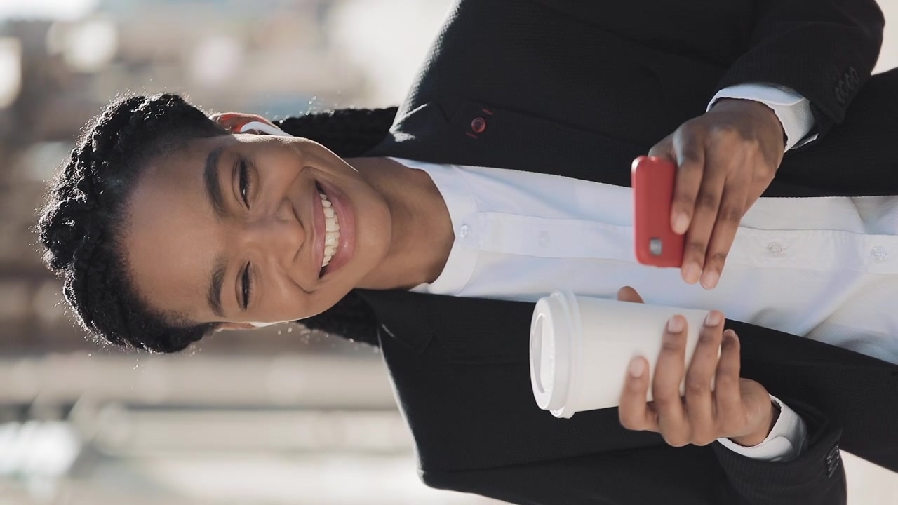 Donna d'affari sorridente che usa lo smartphone e tiene una tazza di caffè all'aperto