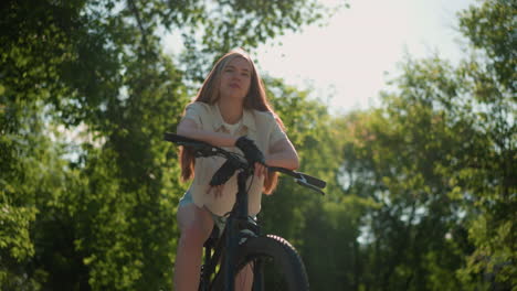 lower angle view of lady leaning on bike handlebar with a subtle smile, sunlight softly illuminating her face, background is filled with vibrant greenery