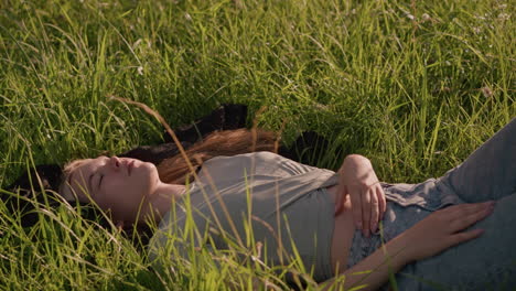 young woman lying in grassy field under warm sunlight, one hand on her leg and other resting on her stomach, immersed in nature, surrounded by tall green grass, she appears calm