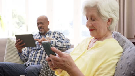 happy diverse senior couple using smartphone and tablet in sunny living room, slow motion