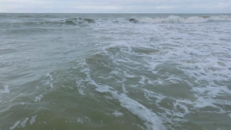 aerial establishing view of big stormy waves breaking against the white sand beach, overcast day, seashore dunes damaged by waves, coastal erosion, climate changes, wide low drone shot moving forward