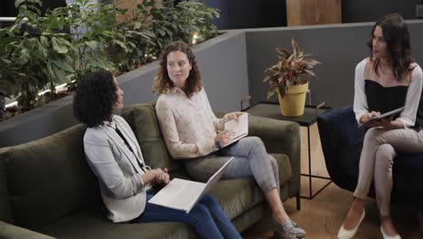 diverse female colleagues in discussion using laptop and tablet in office lounge, slow motion