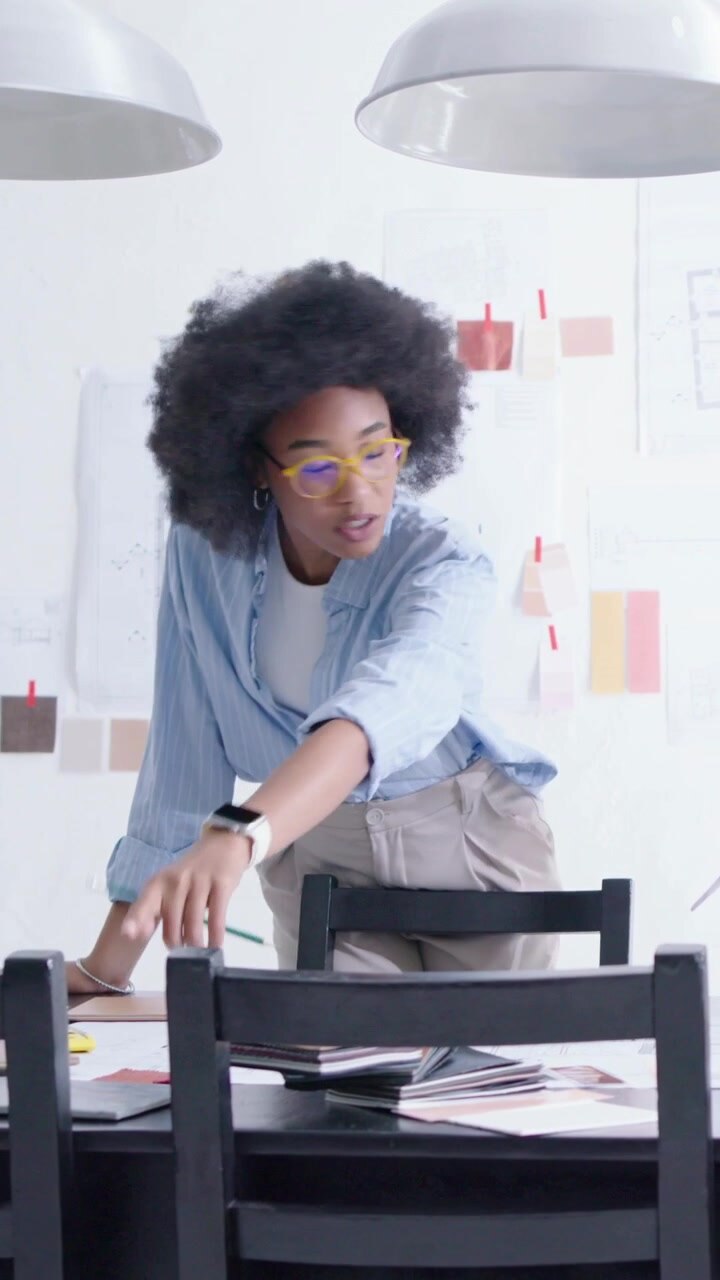 Vertical video: Leaning forward woman with glasses reviewing layouts at studio table with swatches