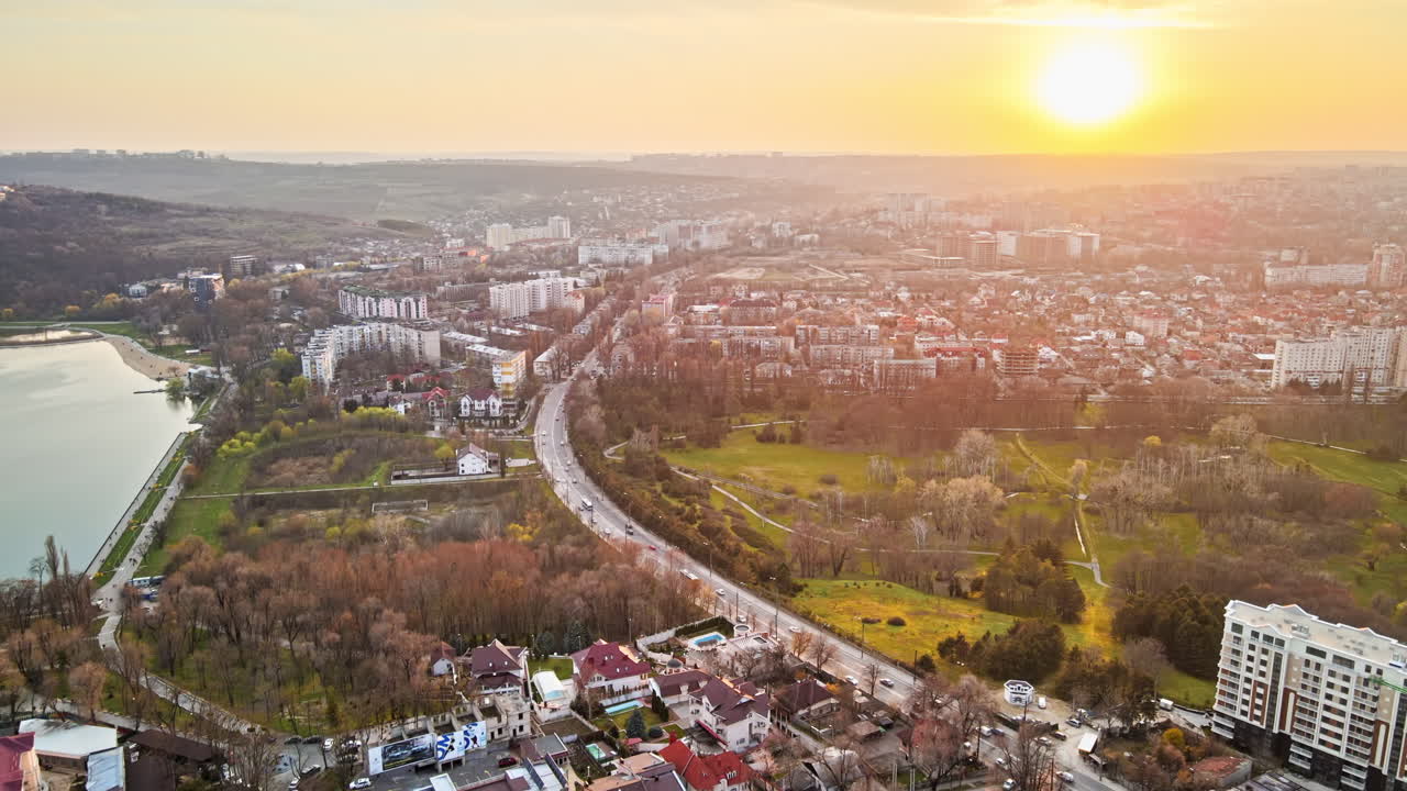 Aerial drone view of Chisinau at sunset. Multiple buildings, trees, park with a lake, road with moving cars, setting sun. Moldova