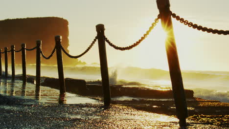 golden hour glow in slow motion as water crashes and sparkles from light against backlit silhouettes of fence posts and chains by the ocean