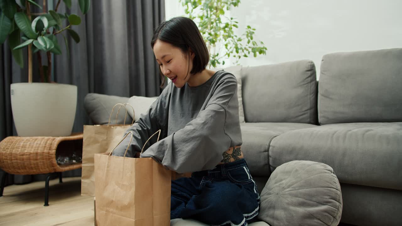 自宅のリビングルームの床に座って、新しい服が入った紙袋を開けているかわいいアジア人女性