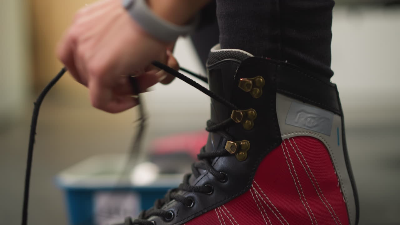 Closeup of lady tying laces on red black ice skates with focus on golden hooks and hand pulling lace, preparing for ice skating activity inside locker room with blurred background creating detail emphasis