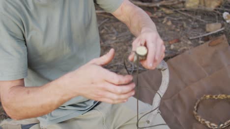 Caucasian-male-survivalist-tying-cord-to-branch-to-make-a-fire-bow-at-camp-in-wilderness