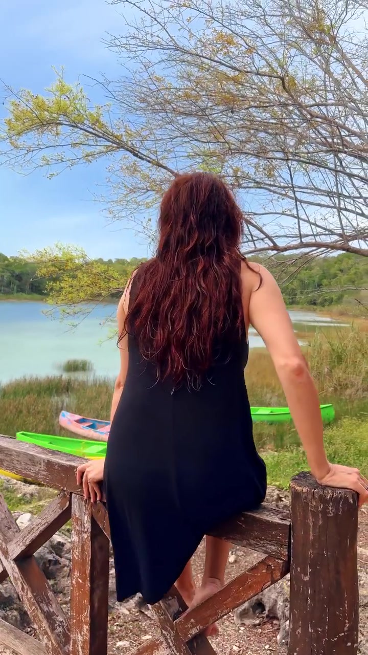 Woman sits on a railing by the water in a natural setting with kayaks nearby. A woman sits on a wooden railing by the water