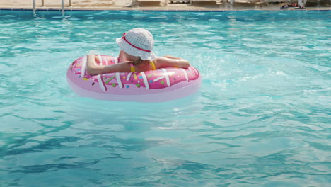 a funny little girl in a panama skates on an inflatable circle in a large pool in a hotel on the sea