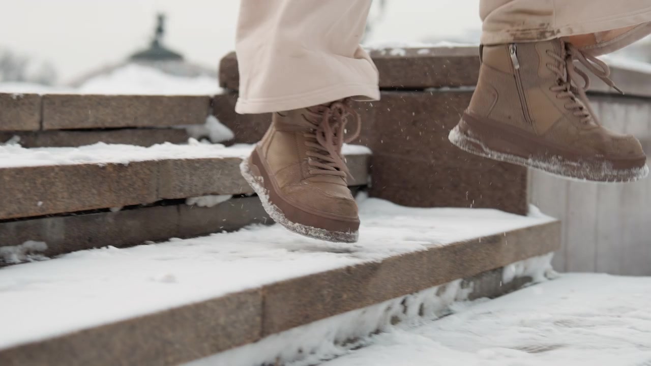 Low-angle close view of person moving on snow-dusted stone steps in overcast daylight, showing brown textured winter boots and beige trousers captured through sequence of actions including stepping