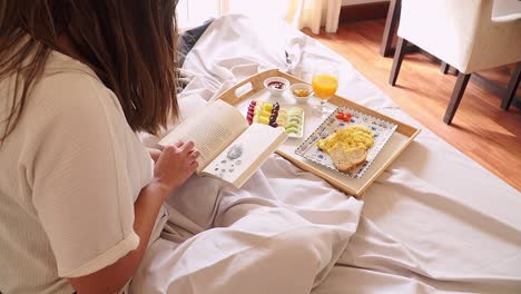 Mujer-Joven-Relajada-Leyendo-Un-Libro-Con-Desayuno-En-Una-Cama-De-Habitación-De-Hotel-De-Vacaciones