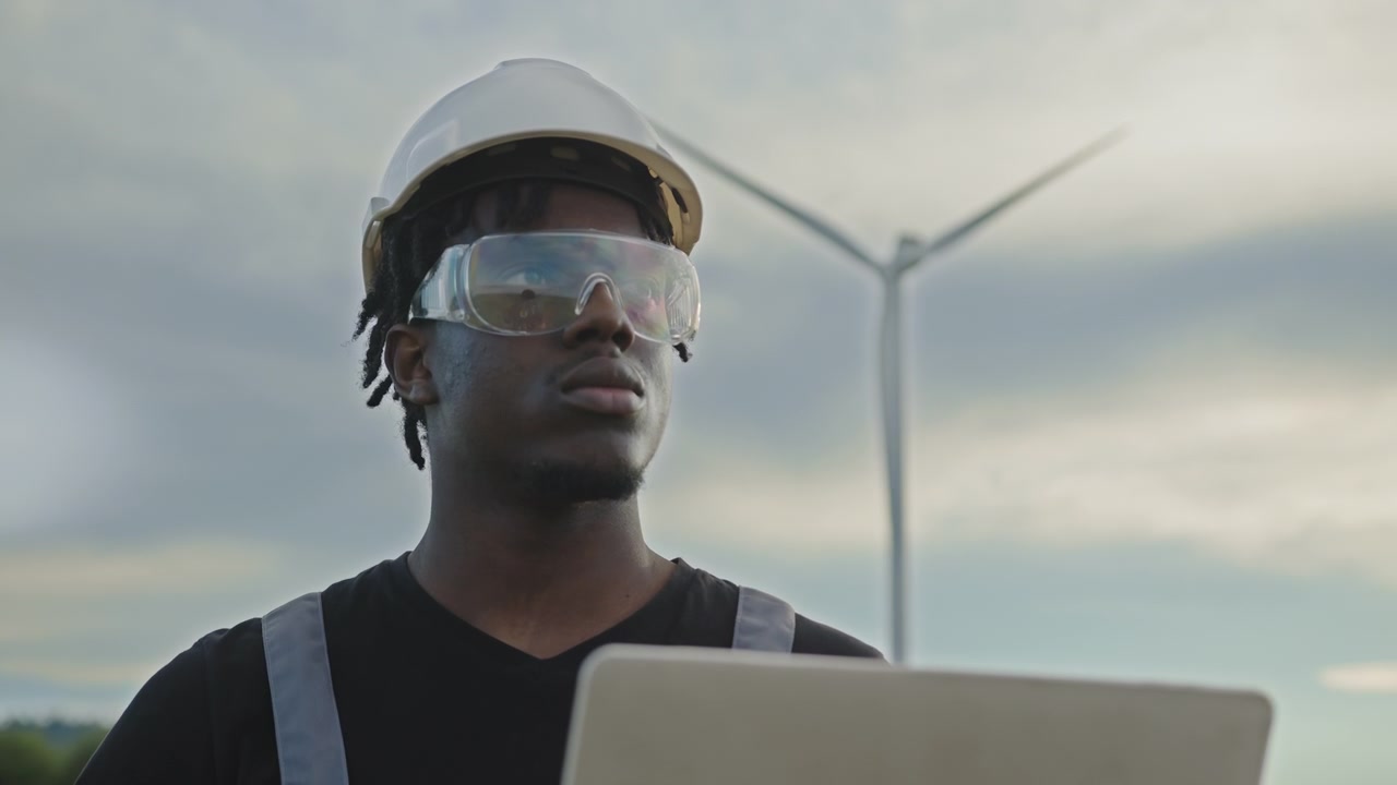 Optagelser viser en afroamerikansk vindmølleingeniør, der arbejder på en bærbar computer foran vindmøller.