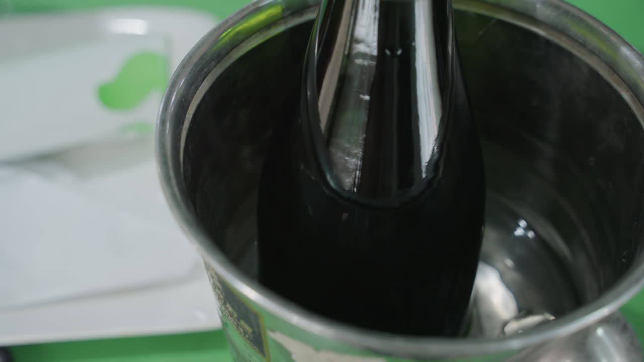 Primo piano di una bottiglia nera che si raffredda in un secchiello, contenitore in acciaio inossidabile mezzo pieno d'acqua e ghiaccio, collo scuro della bottiglia che si innalza con condensa, superficie riflettente che cattura lo sfondo verde, intimo