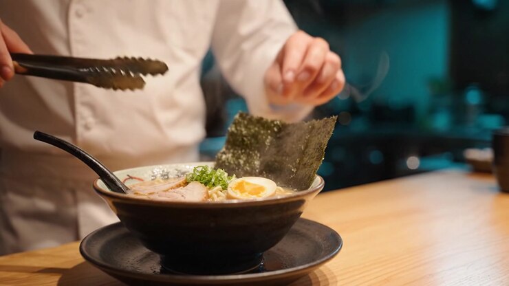 Uno chef aggiunge con maestria il nori a una ciotola fumante di ramen, mettendo in mostra l'intricata arte della preparazione dei noodle giapponesi in un'accogliente cornice culinaria.