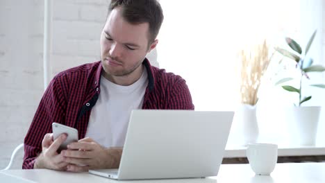 young man using smartphone while working on laptop