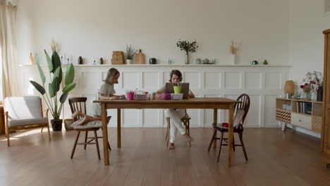 wide shot of a mother working at home on her laptop when her daughter comes in and sits down next to her to do her homework