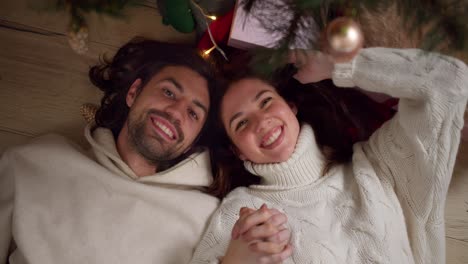 Portrait-of-a-guy-and-a-girl-in-white-sweaters-lying-on-the-floor-near-the-New-Year-tree-holding-hands-smiling-and-looking-at-the-camera-in-a-cozy-room-in-New-Year's-winter