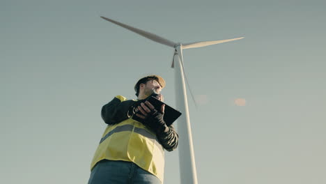 un joven y enfocado ingeniero caucásico con casco blanco usa tecnología en una tableta para auditar turbinas eólicas en un día soleado, promoviendo el uso de energía limpia para un futuro sostenible