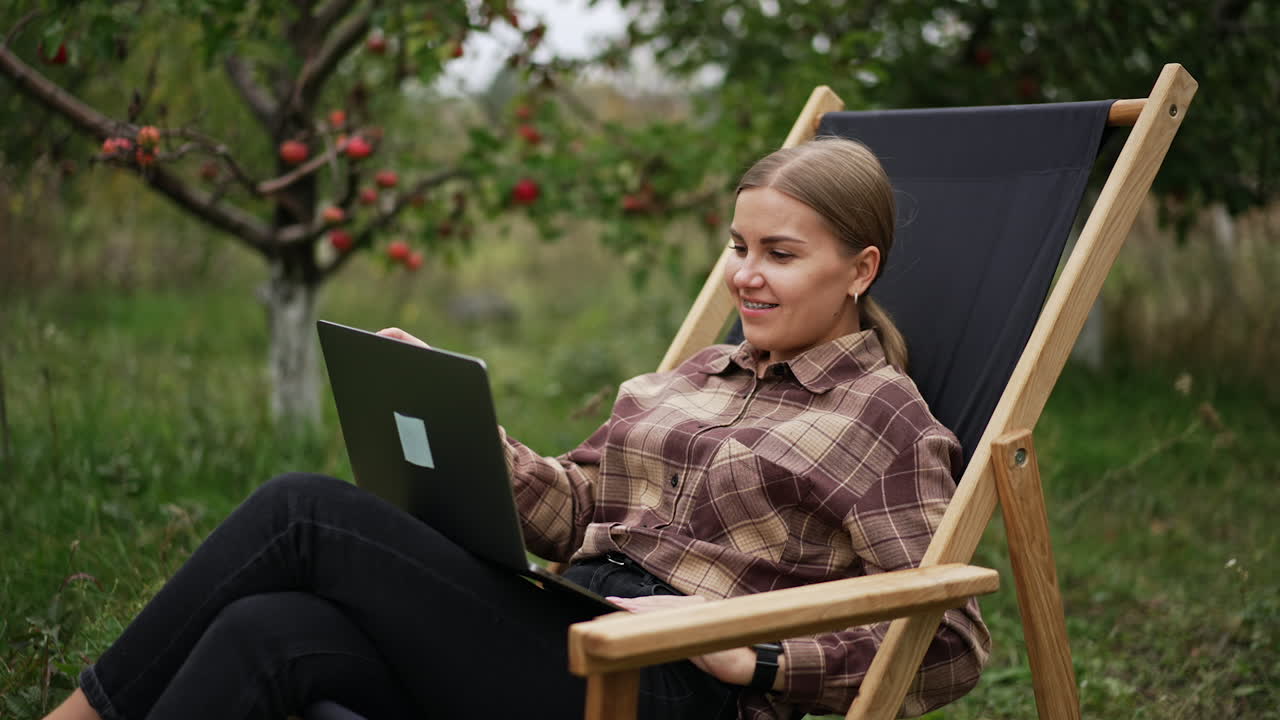 Calm Caucasian woman in checkered shirt sits in chair outdoors. Freelancing lady typing message on the laptop on her laps.