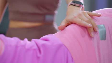 primer plano de una mujer de marrón con un reloj inteligente colocando su mano en la cintura de otra mujer de rosa, acostada durante una sesión de entrenamiento enfocada en un gimnasio