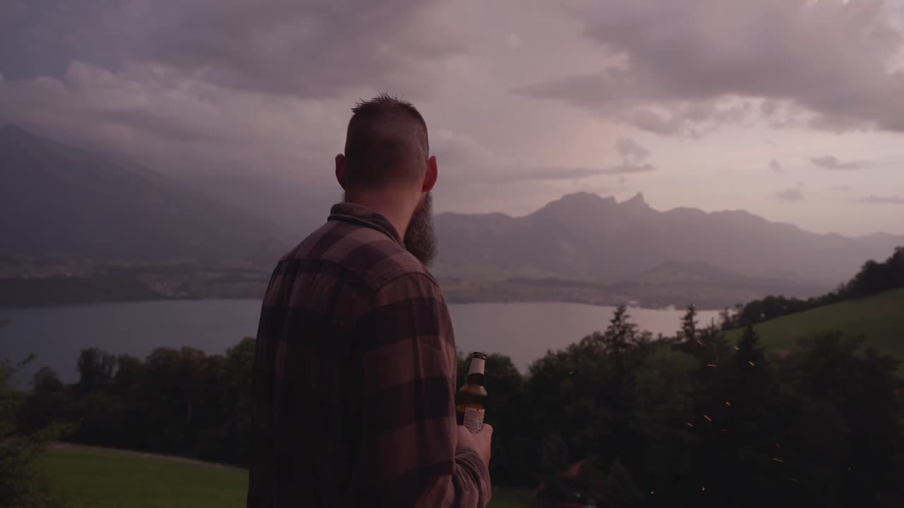 A man stands near a burning campfire on a grassy hill with mountains and a lake in the background at sunset. Concept of solitude, reflection, wilderness adventure, and peaceful living