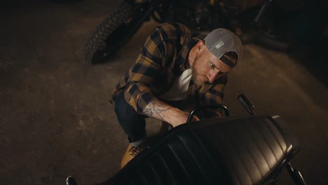 top view of a guy mechanic in a plaid shirt and cap repairing a motorcycle in his workshop studio