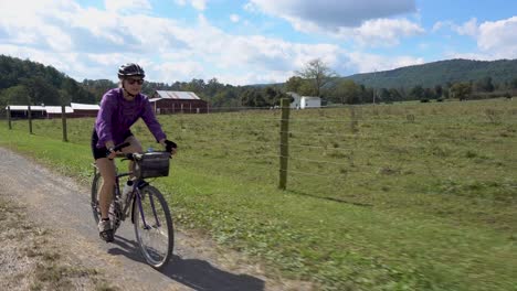 mujer madura con gafas de sol en bicicleta por un camino de ripio con una granja en la distancia