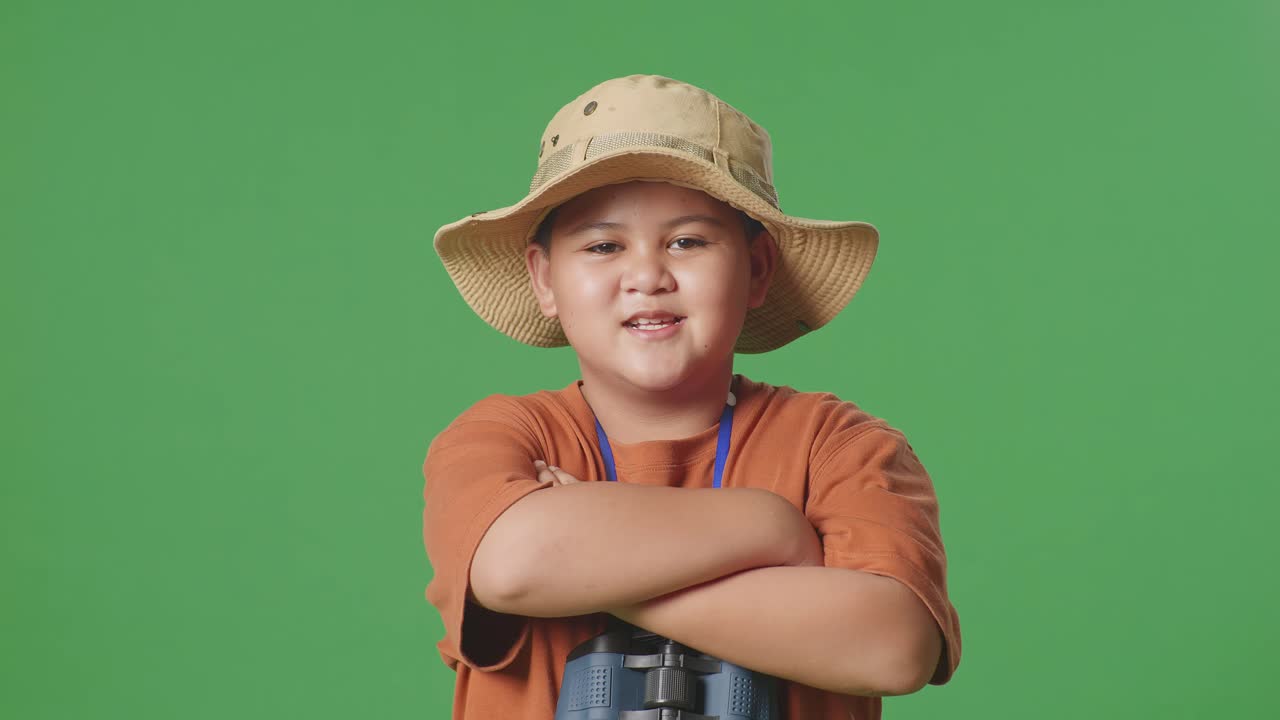 모자와 안경을 쓴 아시아 관광 소년은 녹색 스크린 배경에 서서 팔을 고 카메라에 미소 짓고 있습니다. 소년 연구자는 여행 모험 개념, 근접을 조사합니다.
