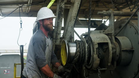 mid-aged bearded male wearing a helmet stands at the working machine rolling the rope. worker of the site for oil boring.