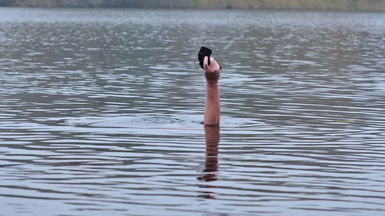 Man in freezing water, leaving only his arm with black hat visible above, 4K