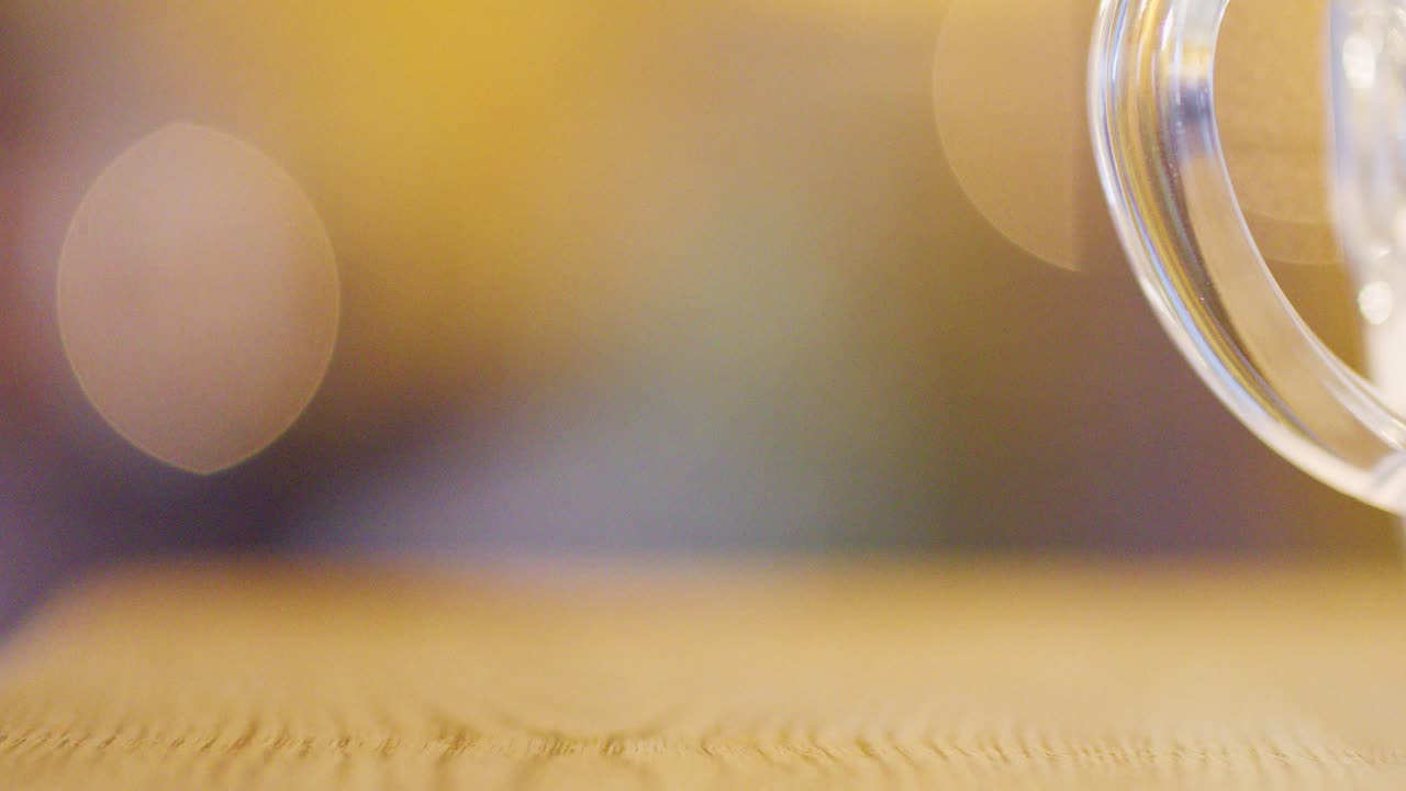 A clear glass sits on a wooden table, surrounded by soft, warm, out-of-focus background lights. Shallow depth of field and gentle ambience