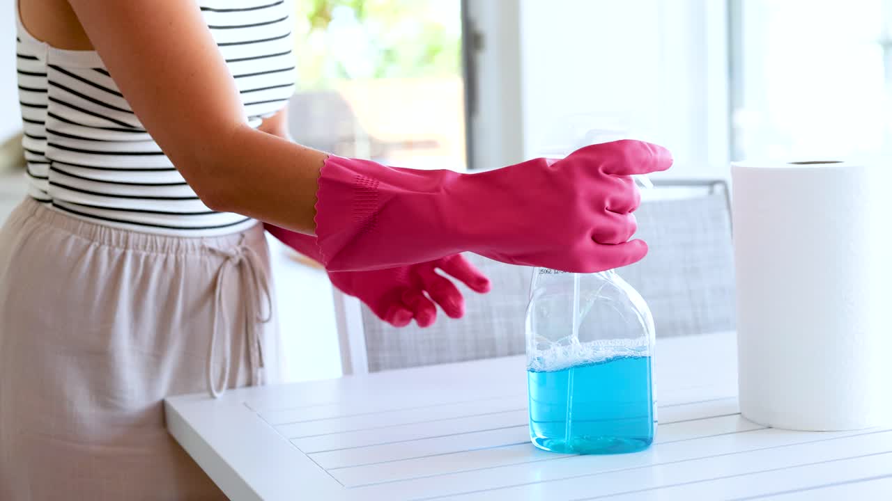 Person wearing gloves cleans a surface using blue spray and paper towel in a bright room