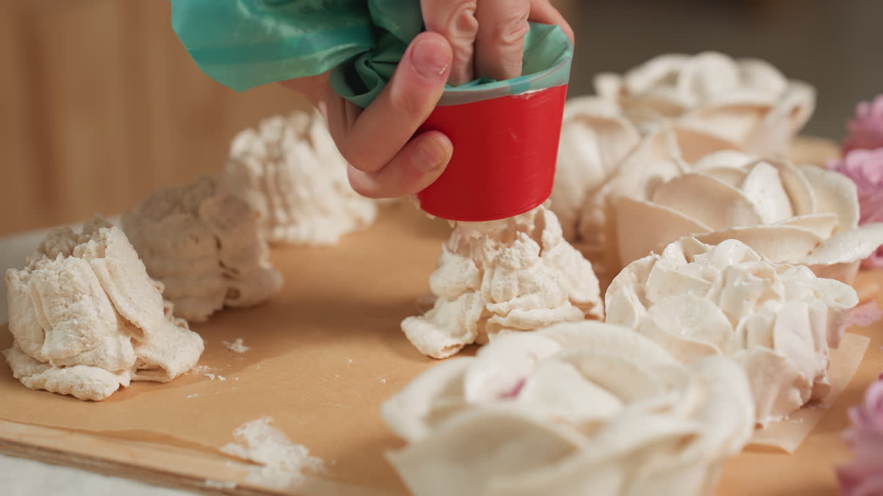 Close up of caterer pressing piping bag with red nozzle while decorative dessert dough drops onto parchment paper, surrounded by flower-shaped frosting cakes and unfinished piped designs