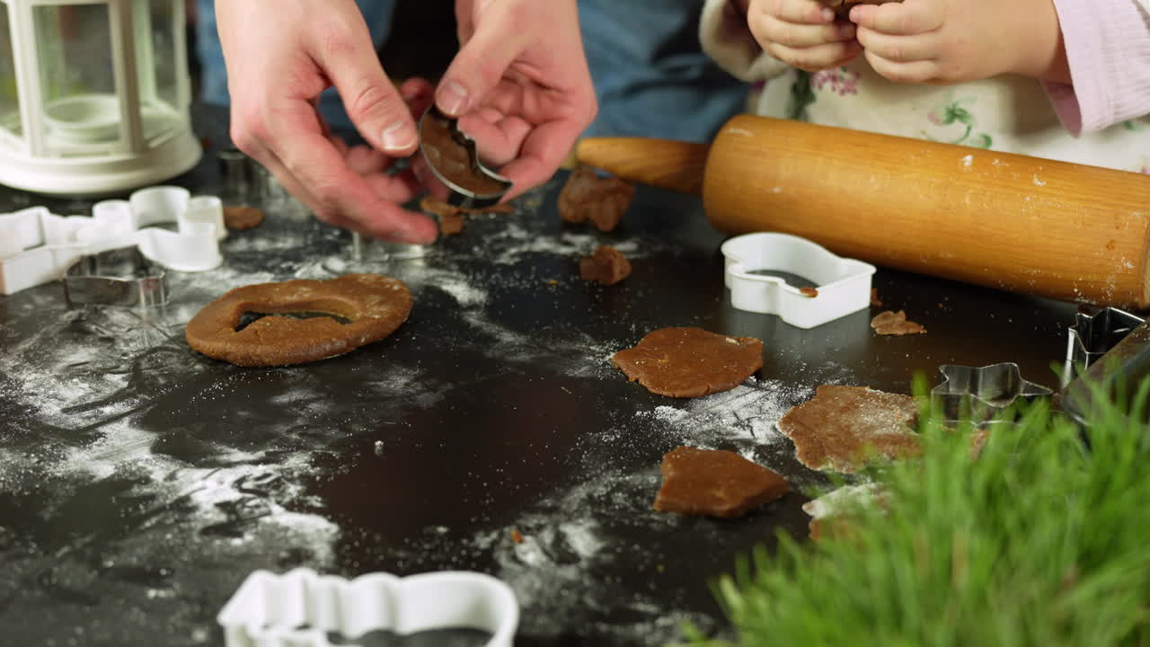 Mother and daughter using cookie cutters to create Christmas gingerbread cookies on a table dusted with flour, surrounded by cookie cutters, rolling pin, and festive decorations