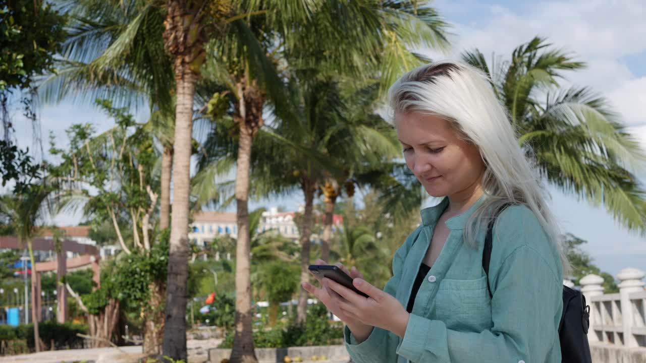 hermosa mujer delgada con el cabello largo rubio en camisa verde de pie cerca de la palmera y el uso de teléfono inteligente sobre el fondo del parque. niña en la pantalla cuadrada tocando y sonriendo