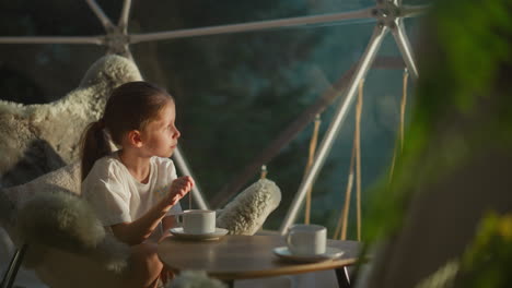 child stirs sweet tea sitting at table. peaceful girl looks at beautiful landscape behind transparent wall of glamping tent. happy vacation