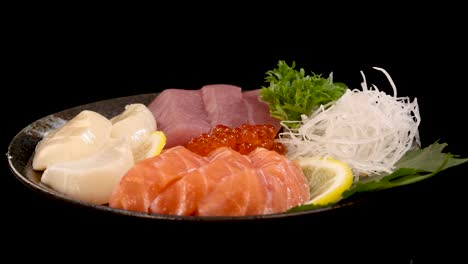a plate of assorted sashimi, including salmon, tuna, scallop, and salmon roe, rotates smoothly under bright studio lighting against a seamless black background