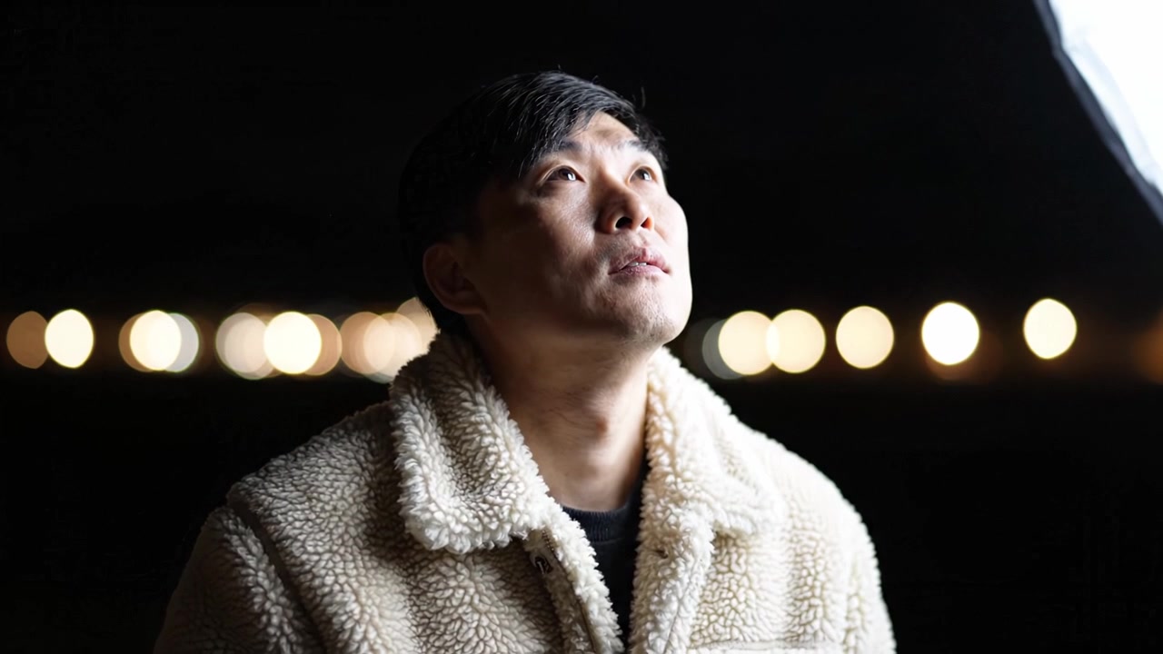 夜空の下、遠くの光を見つめ思索にふける男性。表情豊かな顔にソフトフォーカス。