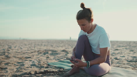 sportswoman sitting on yoga mat checking her smartphone on the beach.