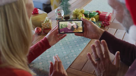 pareja caucásica con sombreros de santa usando un teléfono inteligente para una videollamada de navidad con la pareja en la pantalla
