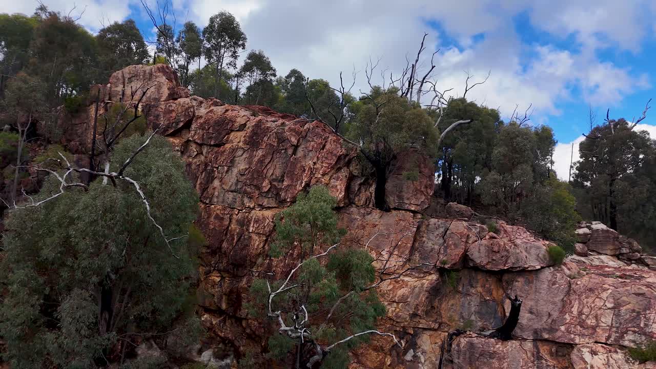 Drone camera glides over rugged sandstone cliffs and eucalyptus trees under partly cloudy skies in Warrumbungle National Park, revealing dramatic Australian bushland terrain