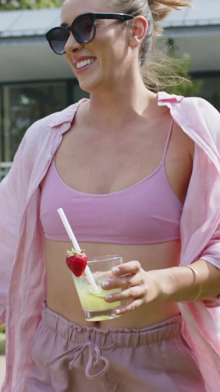 Video verticale: Donna che ride in reggiseno rosa con occhiali da sole che saluta un'amica in giardino, tenendo un bicchiere.