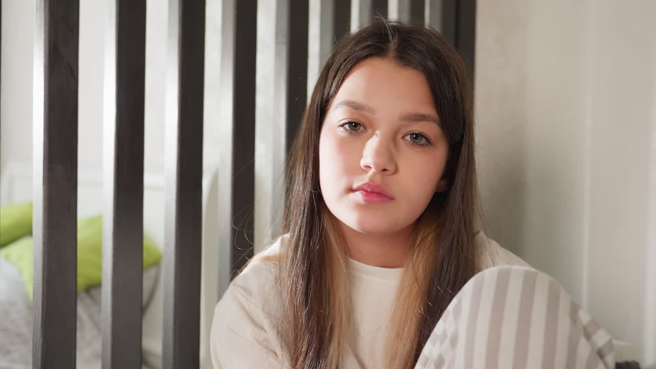 Zoom in on holiday kid seated calmly in striped night wear, long hair draped, cozy bedroom with partition rails, bed pillows behind, relaxed thoughtful gaze during quiet home moment