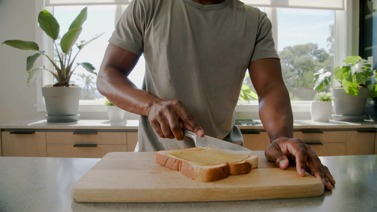 Placing smeared knife, man in gray tee spreading spread on bread at kitchen counter making snack